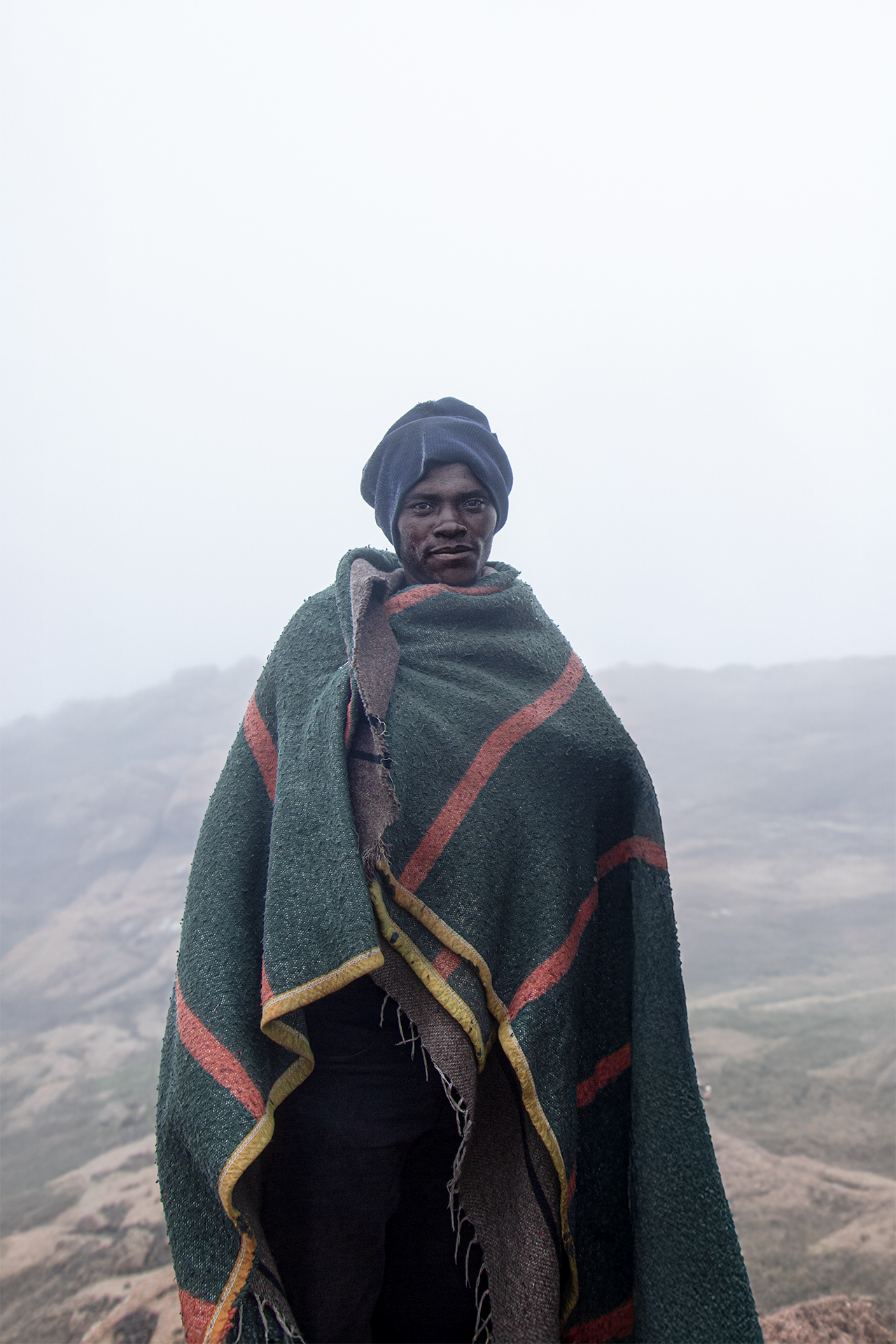 Portrait of a shepherd standing in the misty highlands of Thabana Ntlenyana, the highest peak in Lesotho. Wrapped in a thick woolen blanket, he faces the cold mountain air with resilience, embodying the traditional lifestyle of Basotho herders who endure harsh conditions in the high-altitude pastures.